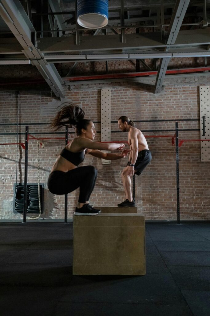A woman and a man do box jumps in a brick-walled gym. The woman is in mid-air, and the man stands on his box.