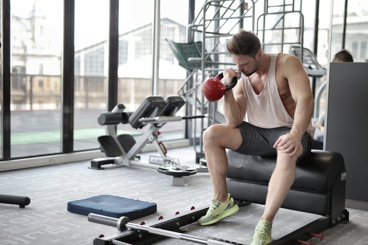 A man in a tank top sits on a gym bench, holding a red kettlebell to his chin. He wears grey shorts and neon sneakers.