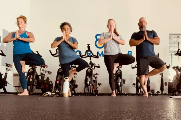 Three women and a man doing the tree pose (vrikshasana) in front of exercise bicycles.