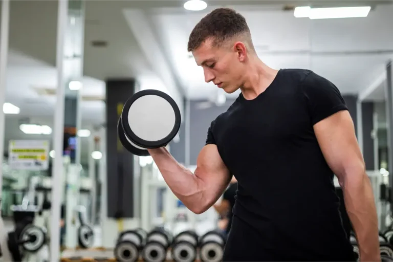A man in a black t-shirt lifts a dumbbell with his right arm at a gym.