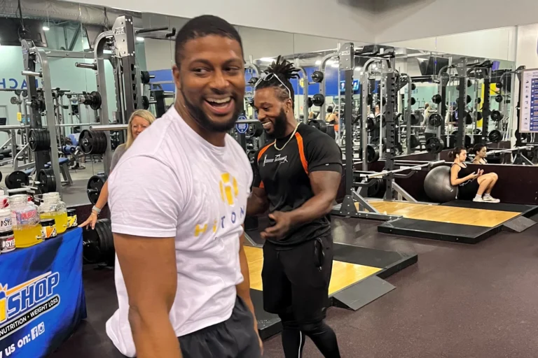 A smiling man in the foreground is at the gym with mirrors and fitness equipment. Another man and a woman smile behind him.