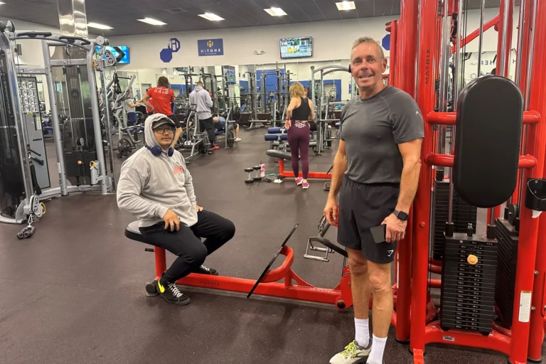 One man is standing in front of the seated row machine, while another is sitting on it.