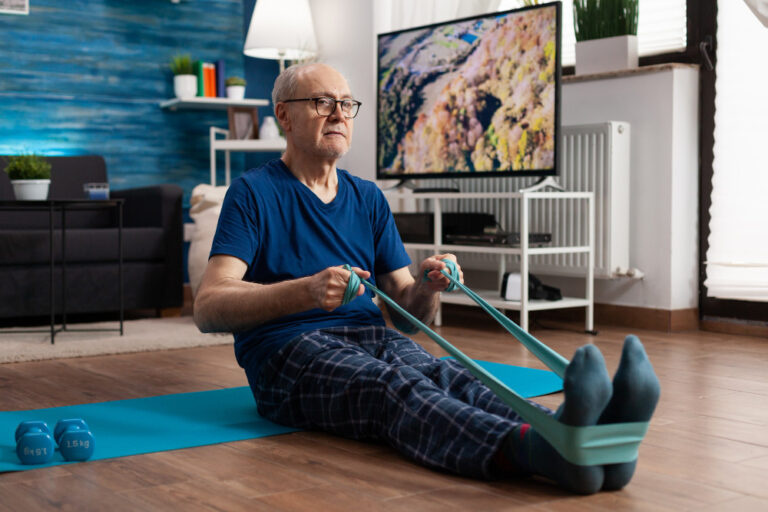 A senior man sits on the living room floor with feet straight and pulls on a resistance band that goes around his feet.