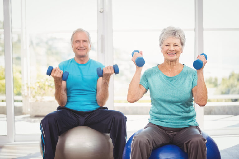 An elderly man and woman perform seated exercises with dumbbells on stability balls, smiling and wearing workout clothes.