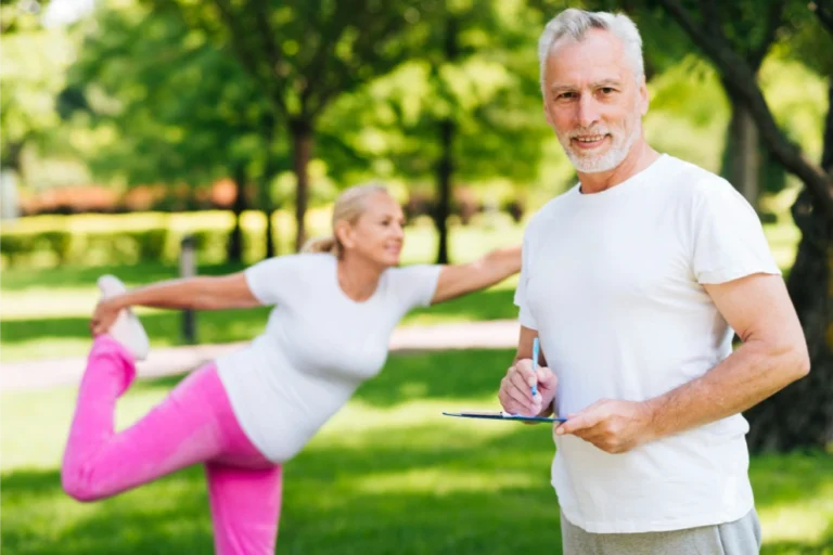 A man in a white t-shirt holds a clipboard while a woman in pink pants and a white shirt performs a stretching pose in a park.