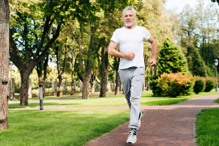 An elderly man in a white t-shirt and gray sweatpants jogs along a path in a park with trees and greenery surrounding him.