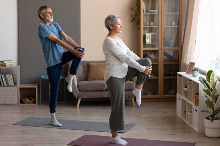 A senior man and woman practice a balance exercise, standing on one leg with arms raised, in a well-lit living room.