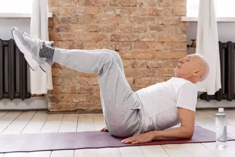An elderly man in a white shirt and gray sweatpants performs leg raises on a yoga mat with a water bottle nearby.