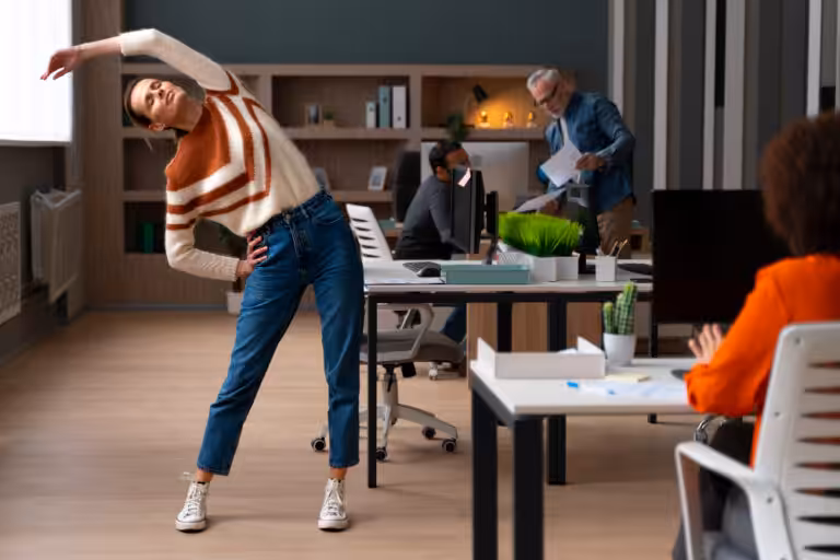 A woman stretches to the side in an office, with two colleagues in the background and one in the foreground working.