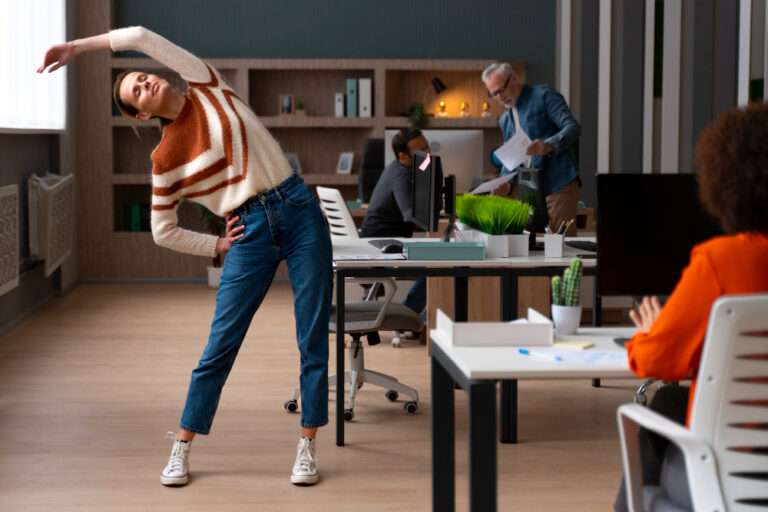 A woman stretches to the side in an office, with two colleagues in the background and one in the foreground working.