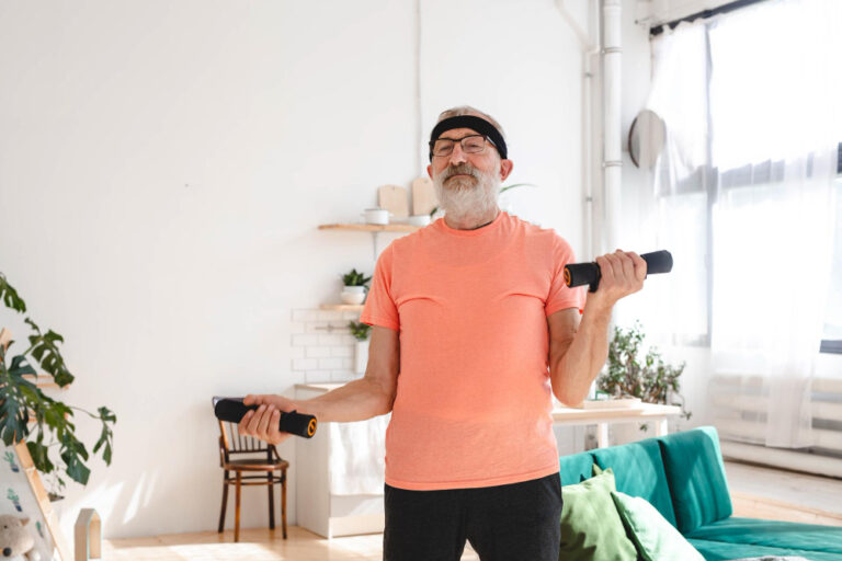 An elderly man in an orange t-shirt and headband holds weights in both hands while standing in a well-lit room.
