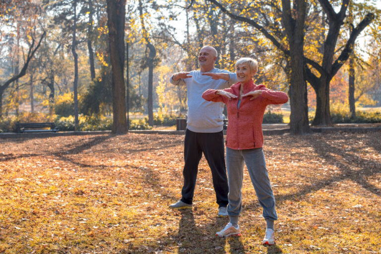 A woman in a red jacket and a man in gray sweatshirt, both in their 60s, smile, stand and stretch arms at a park.