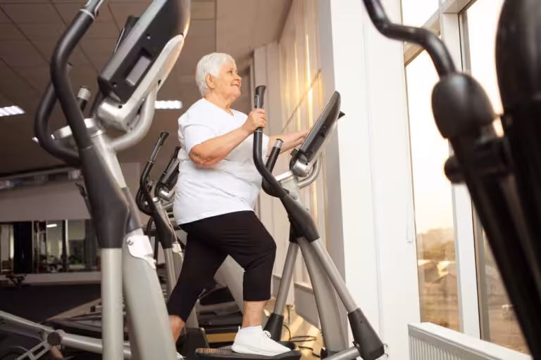 An elderly woman walks on an elliptical machine in a gym, wearing a white t-shirt, black pants, and white sneakers.