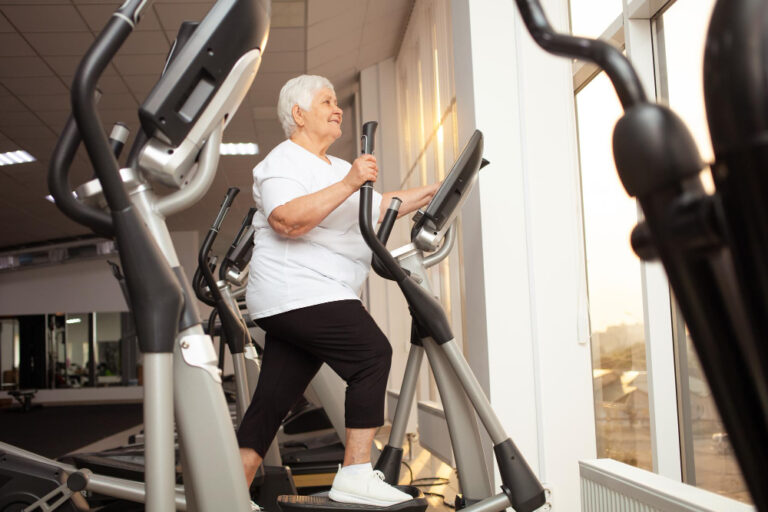 An elderly woman walks on an elliptical machine in a gym, wearing a white t-shirt, black pants, and white sneakers.