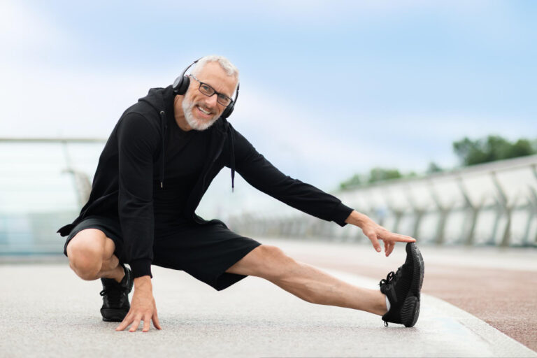 A man in his 50s stretches his leg outdoors, wearing black headphones, sweatshirt, shorts and sneakers.