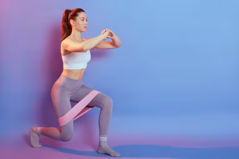 A woman in a white top and grey leggings does a lunge with a pink resistance band around her thighs against a purple wall.