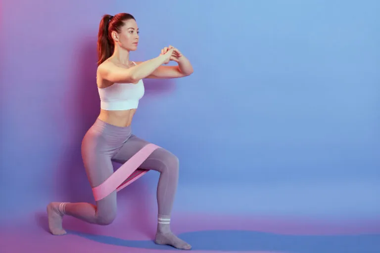 A woman in a white top and grey leggings does a lunge with a pink resistance band around her thighs against a purple wall.