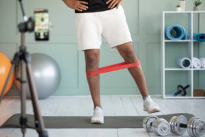 A person in white shorts and shoes uses a red resistance band around their legs, standing on a gray mat with weights nearby.