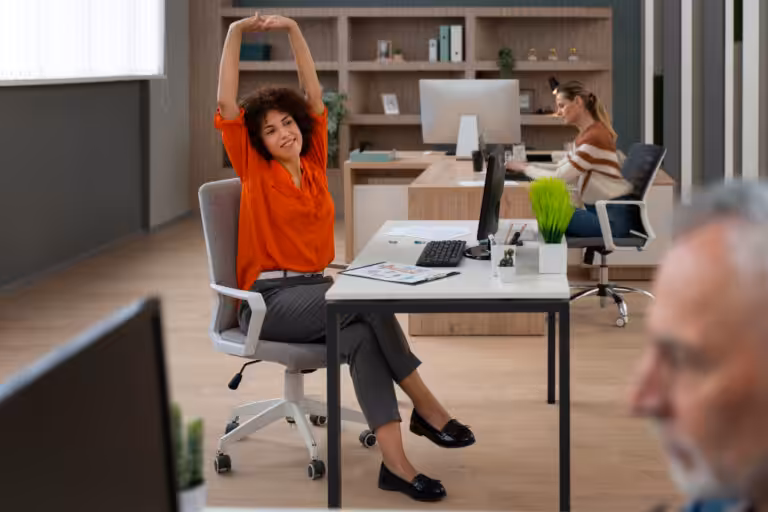 Woman with curly brown hair in an orange shirt stretches arms overhead while seated at a desk in an office.