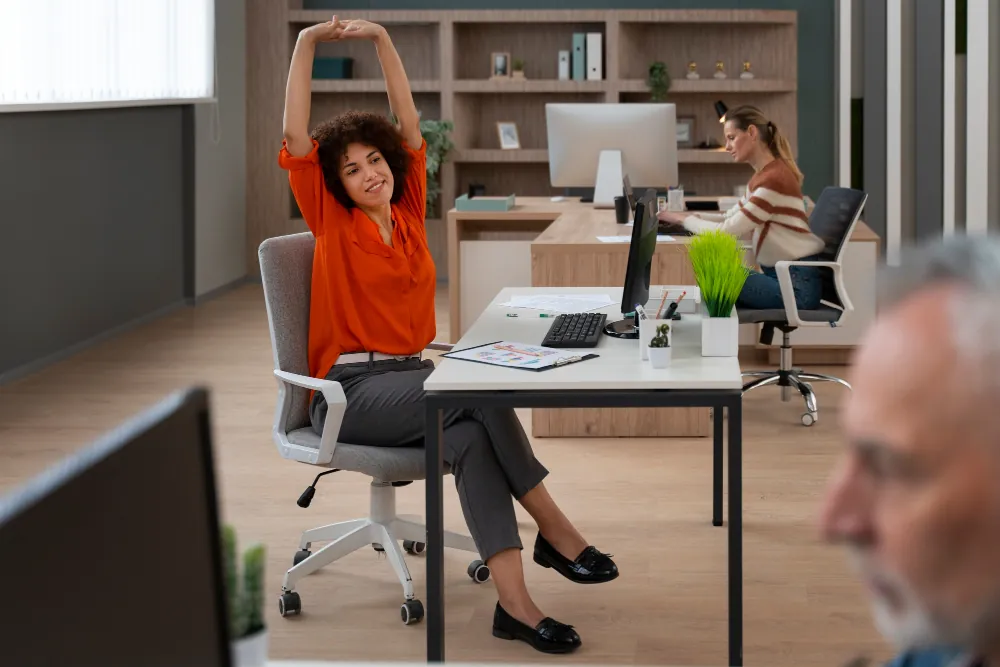 Woman with curly brown hair in an orange shirt stretches arms overhead while seated at a desk in an office.