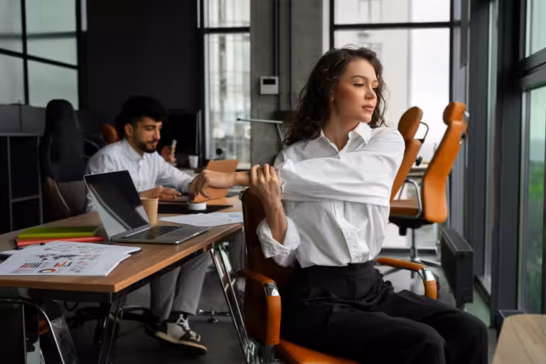 Woman with curly brown hair in a white shirt stretches her arm, sitting in an office; a man works at a desk in the background.
