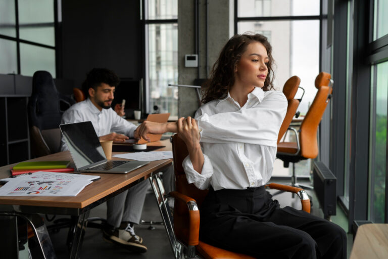 Woman with curly brown hair in a white shirt stretches her arm, sitting in an office; a man works at a desk in the background.