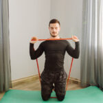 A man in black workout clothes kneels on a green mat, holding a red resistance band, in a room with patterned curtains.