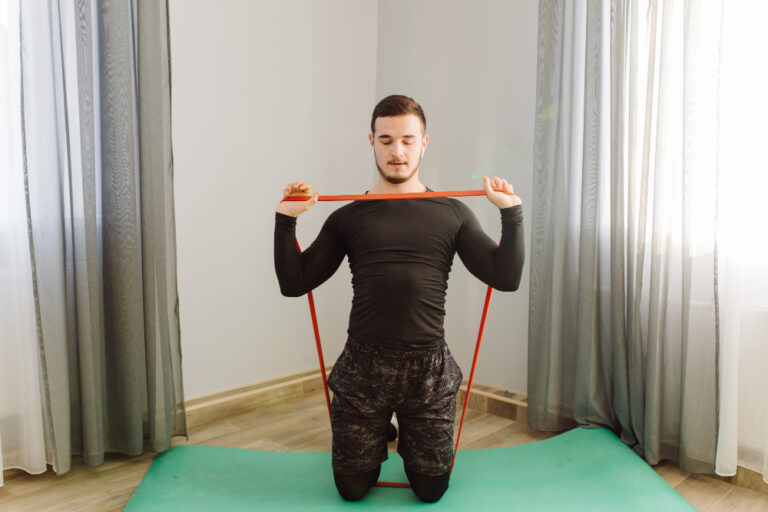 A man in black workout clothes kneels on a green mat, holding a red resistance band, in a room with patterned curtains.
