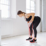 A woman in black workout clothes bends over, holding a red resistance band, in a bright, white room with large windows.