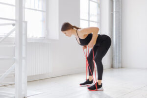 A woman in black workout clothes bends over, holding a red resistance band, in a bright, white room with large windows.