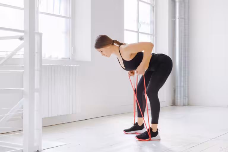 A woman in black workout clothes bends over, holding a red resistance band, in a bright, white room with large windows.