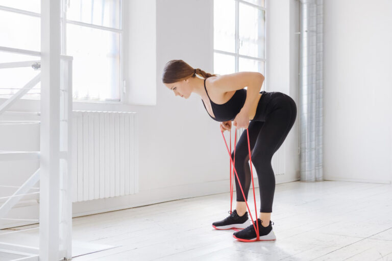 A woman in black workout clothes bends over, holding a red resistance band, in a bright, white room with large windows.