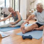 Two older people in workout clothes stretch on mats, a laptop and weights nearby, in a bright room with a couch.