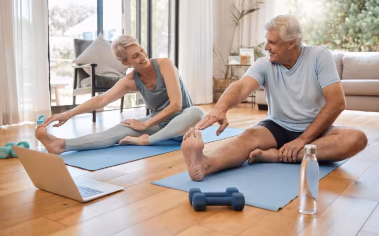 Two older people in workout clothes stretch on mats, a laptop and weights nearby, in a bright room with a couch.