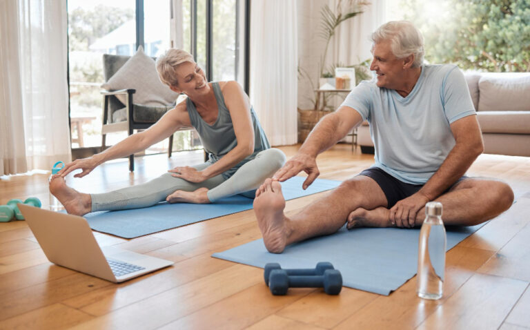 Two older people in workout clothes stretch on mats, a laptop and weights nearby, in a bright room with a couch.
