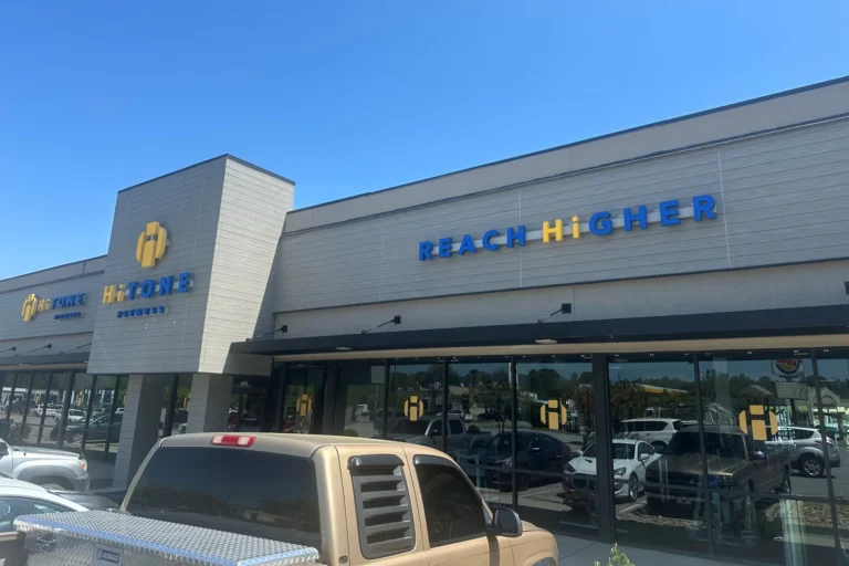 Under a blue sky, a storefront with "reach higher" and "hitone" signage, big windows and parked cars in front.