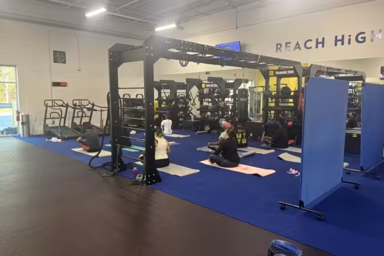Several people sit in a cross-legged pose on mats in a gym with weights, treadmills, and blue flooring.