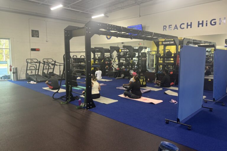 Several people sit in a cross-legged pose on mats in a gym with weights, treadmills, and blue flooring.