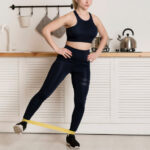 A woman in a dark blue sports bra and leggings does a leg exercise with a yellow resistance band in a kitchen.