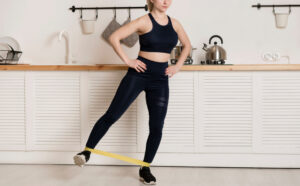 A woman in a dark blue sports bra and leggings does a leg exercise with a yellow resistance band in a kitchen.