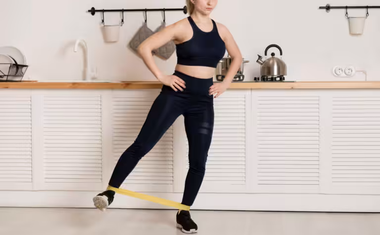 A woman in a dark blue sports bra and leggings does a leg exercise with a yellow resistance band in a kitchen.