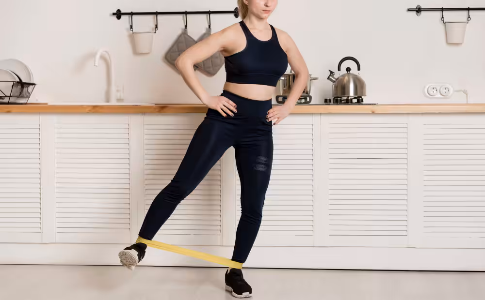 A woman in a dark blue sports bra and leggings does a leg exercise with a yellow resistance band in a kitchen.