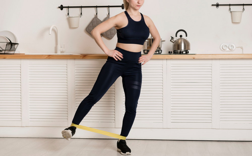 A woman in a dark blue sports bra and leggings does a leg exercise with a yellow resistance band in a kitchen.