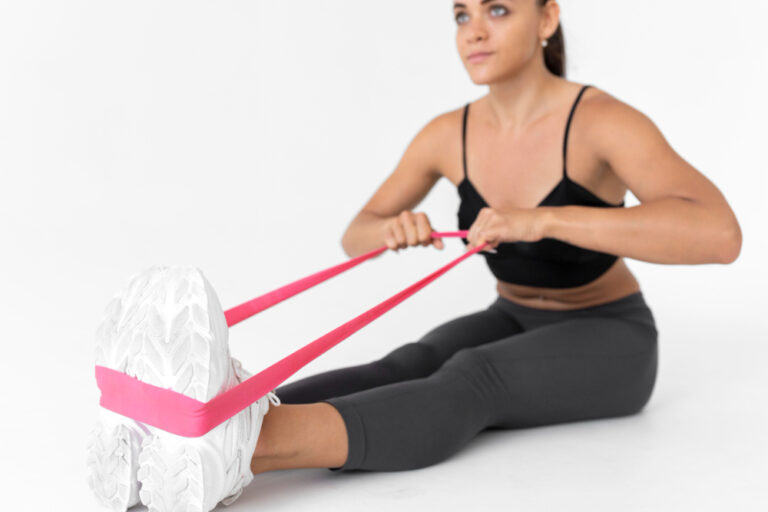 A seated woman in black sportswear pulls a pink resistance band, that's attached to her feet.
