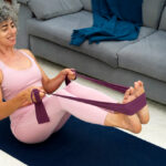 A woman with gray curly hair, in pink sportswear, stretches a purple resistance band with her hands and feet.
