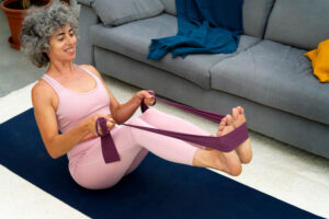 A woman with gray curly hair, in pink sportswear, stretches a purple resistance band with her hands and feet.
