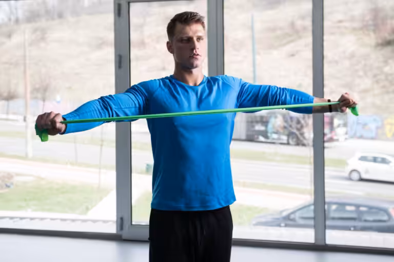 Man with short brown hair in a blue long-sleeved shirt stretches a green resistance band with arms extended.