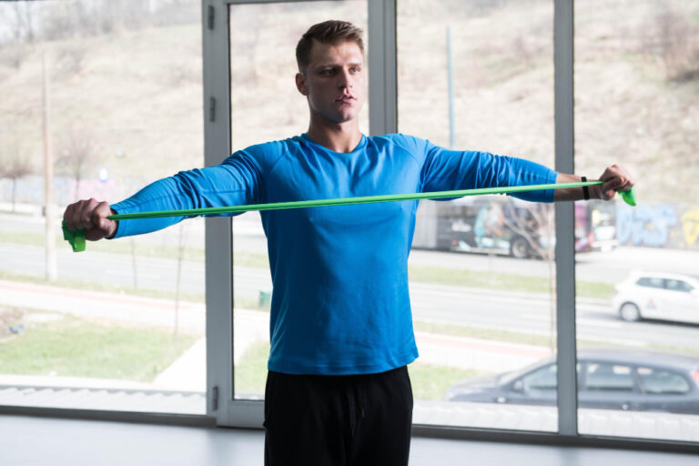 Man with short brown hair in a blue long-sleeved shirt stretches a green resistance band with arms extended.