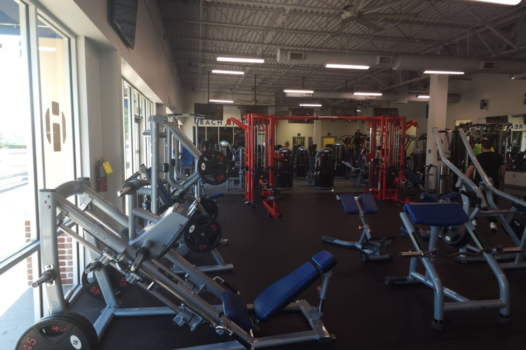 A spacious gym with black flooring and various silver and blue weight machines, and red squat racks.