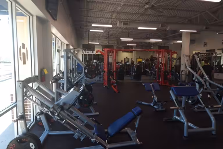 A spacious gym with black flooring and various silver and blue weight machines, and red squat racks.